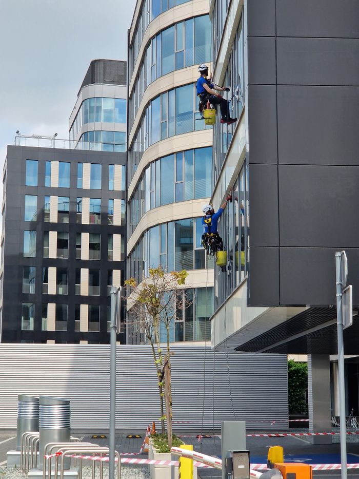 hero-img-01 Deux employés nettoient les fenêtres d’un grand immeuble de bureaux en milieu urbain.